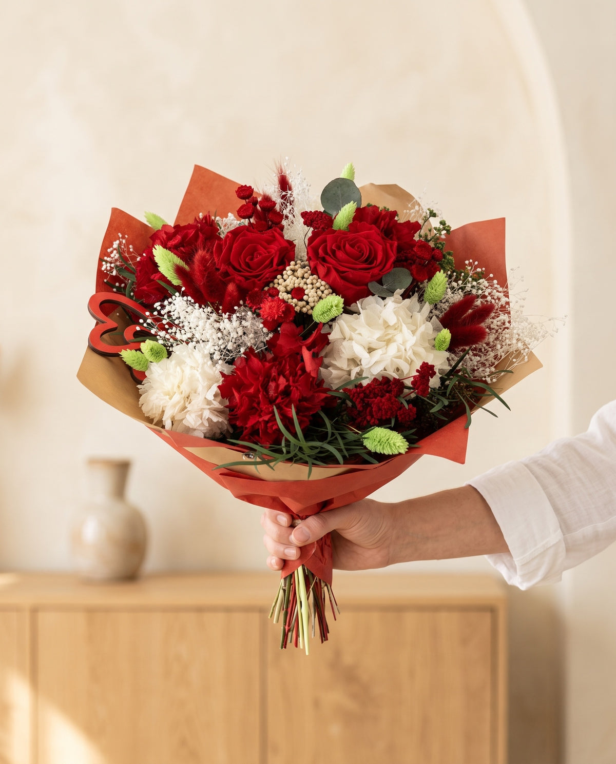 Mano sujetando el ramo de flores secas y preservadas en rojo con rosas con su papel, como se entrega.