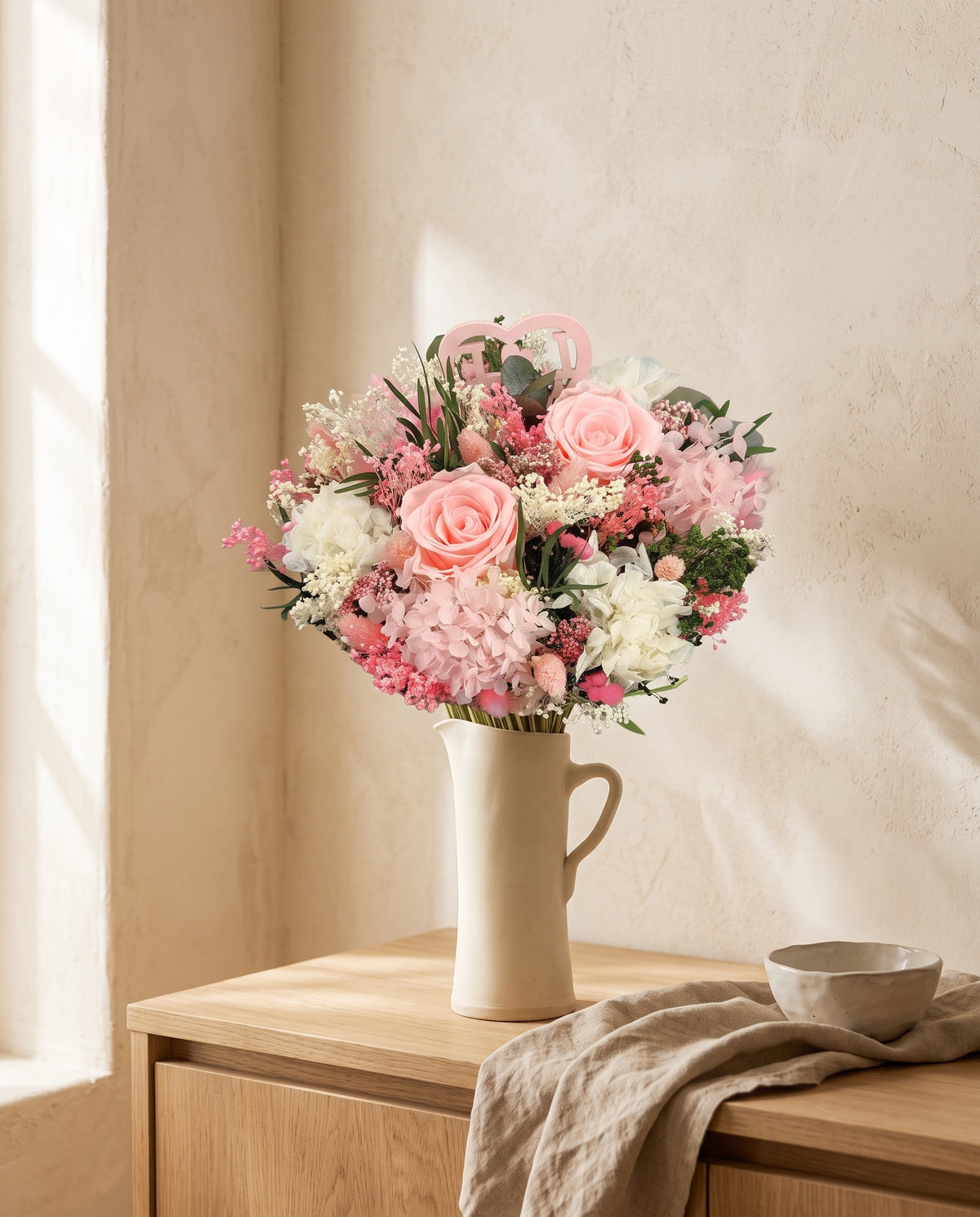 Ramo de flores secas y preservadas en rosa con rosas en una jarra sobre un aparador, ambiente de casa. Jarra no incluida.