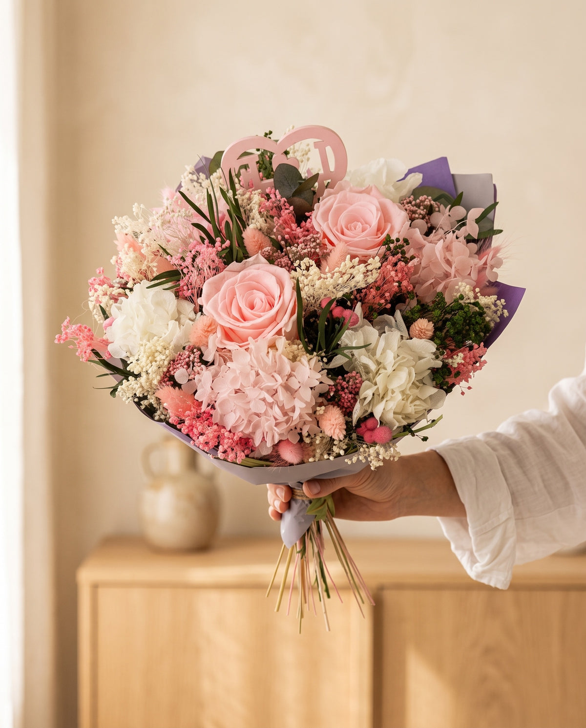 Mano sujetando el ramo de flores secas y preservadas en rosa con rosas con su papel, como se entrega.
