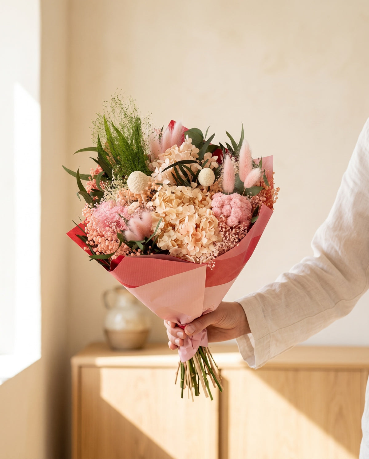 Mano sujetando el ramo de flores secas y preservadas en rosa claro con su papel, como se entrega.