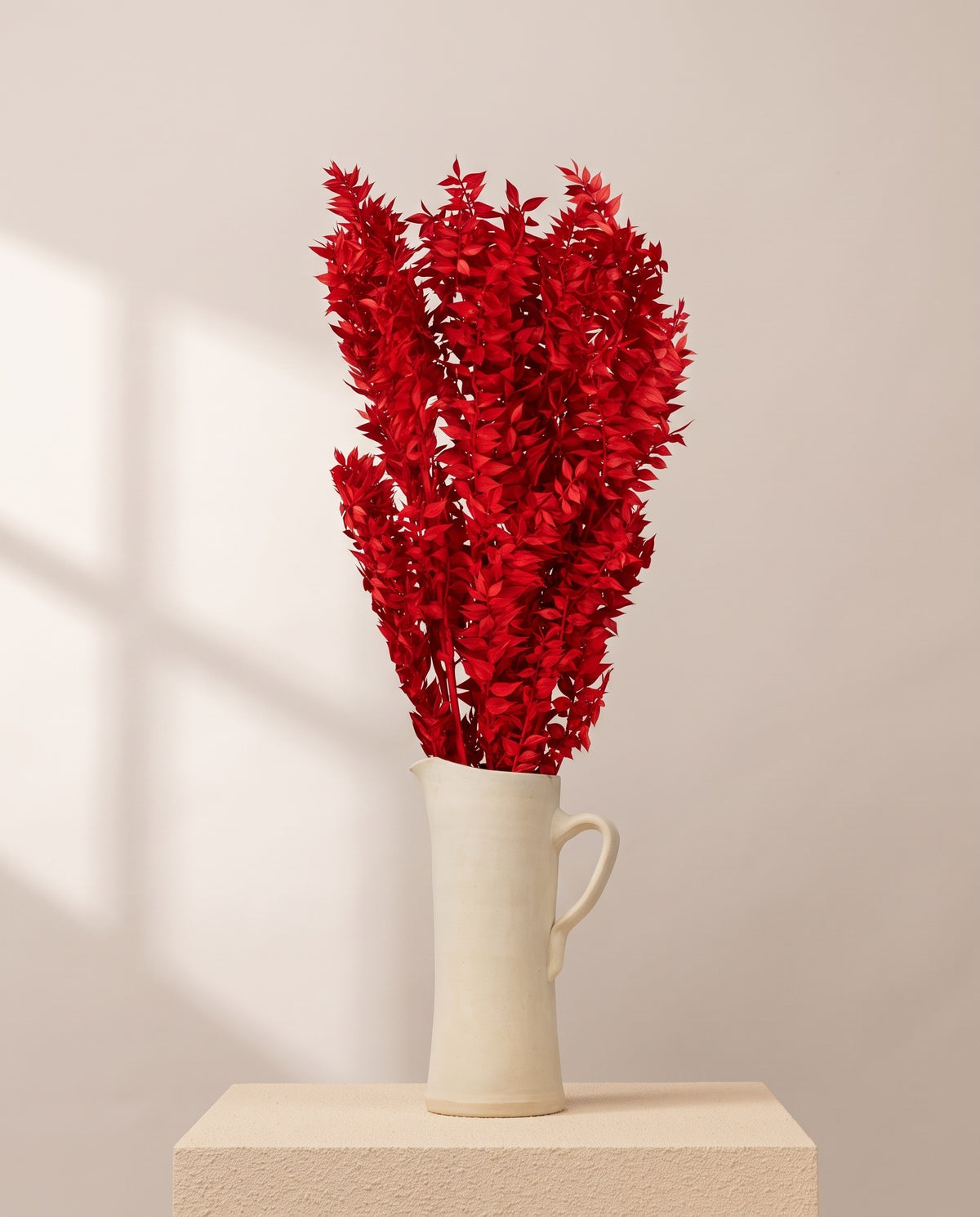 Ruscus preservado rojo en una jarra de cerámica crema, ambientado. Jarra no incluida.